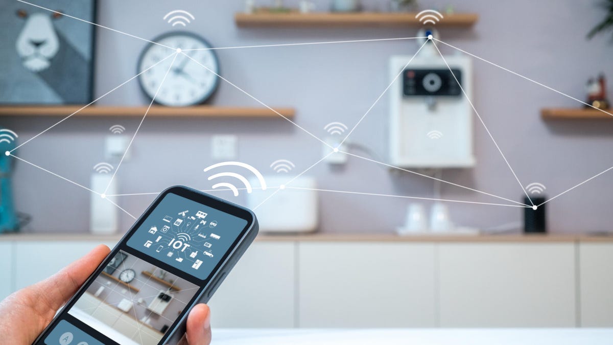 Photo-illustration shows a hand holding a smartphone that displays an app that connects various gadgets in the background (a clock, a speaker and so on). The connections are represented by lines and Wi-Fi symbols.