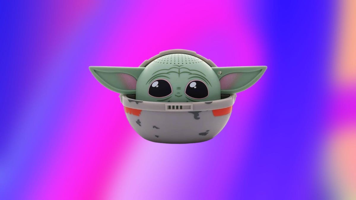 A beige Bluetooth speaker with the head of Grogu peeking out from the top of it, set against a blue and pink background