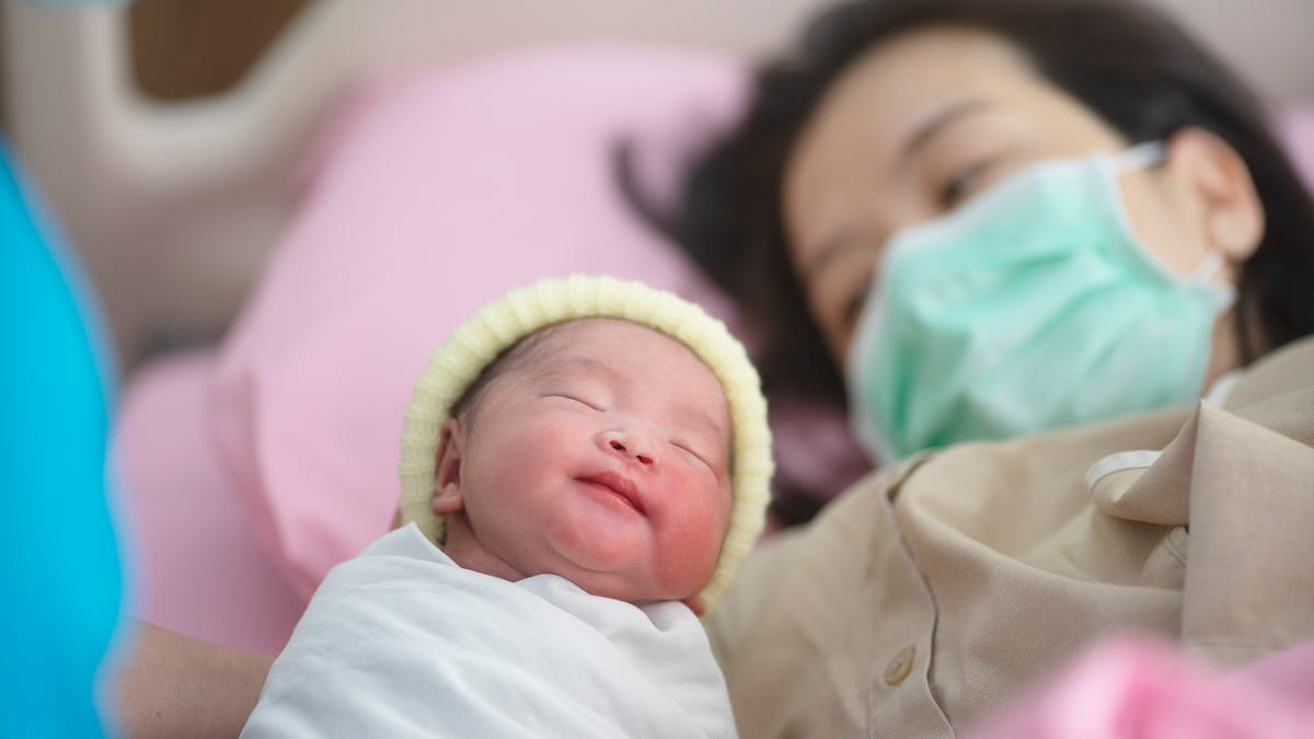 A newborn baby is held up to their mother wearing a face mask.