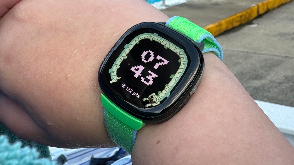 Wrist of a kid wearing Fitbit's new fitness watch, showing a bright display and green straps.