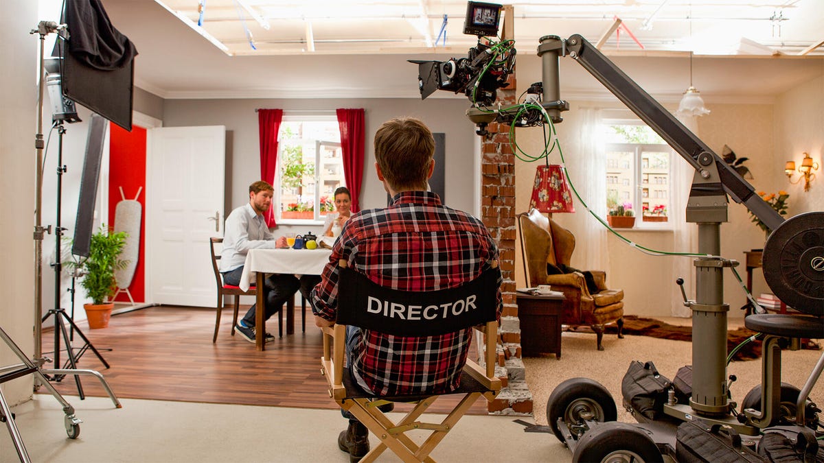 A director sits in a director's chair on set. A camera looms above and two actors sit at a table.
