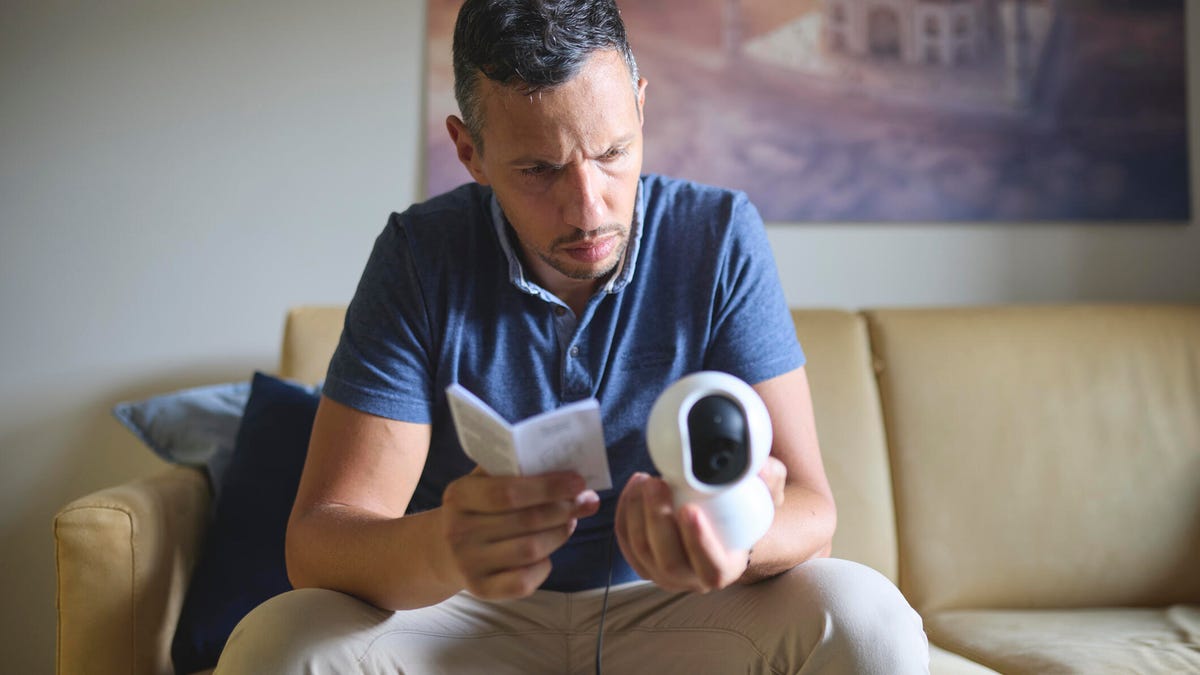 A man on a couch holds a manual and security camera while frowning.