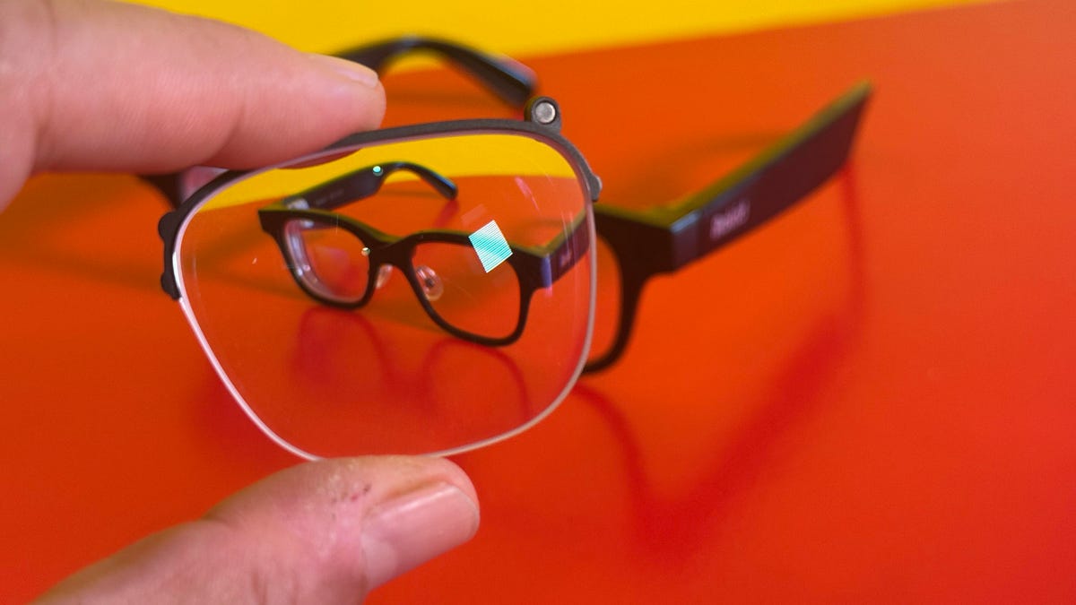 A hand holding up a prescription lens insert in front of a pair of Rokid Glasses