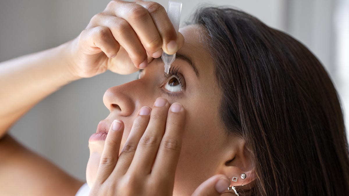 A person with long dark brown hair and silver hoop earrings applying eye drops into their left eye.