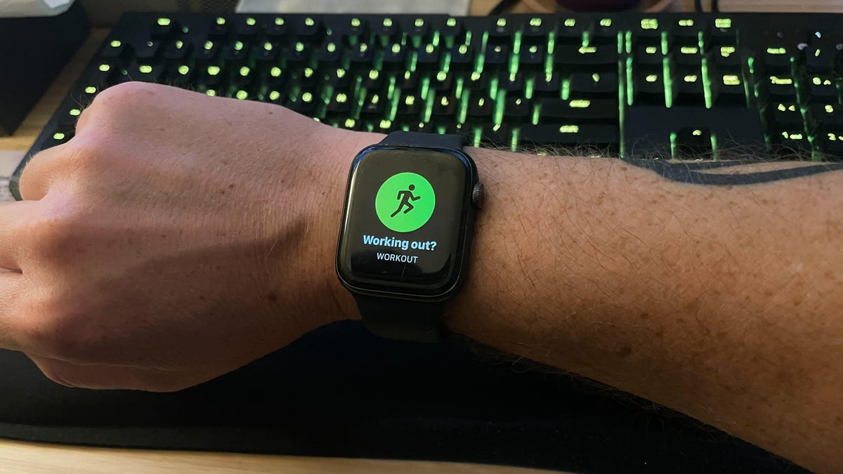 An Apple Watch Series 5 on a wrist with a "Working out?" prompt asking if the user, clearly sitting in front of a keyboard, is currently running.