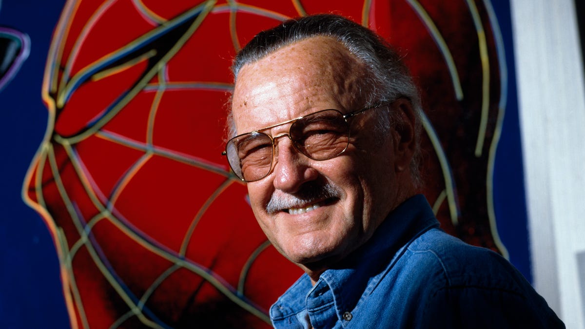 Stan Lee, in trademark moustache and sunglasses, grins in front of a portrait of Spider-Man.
