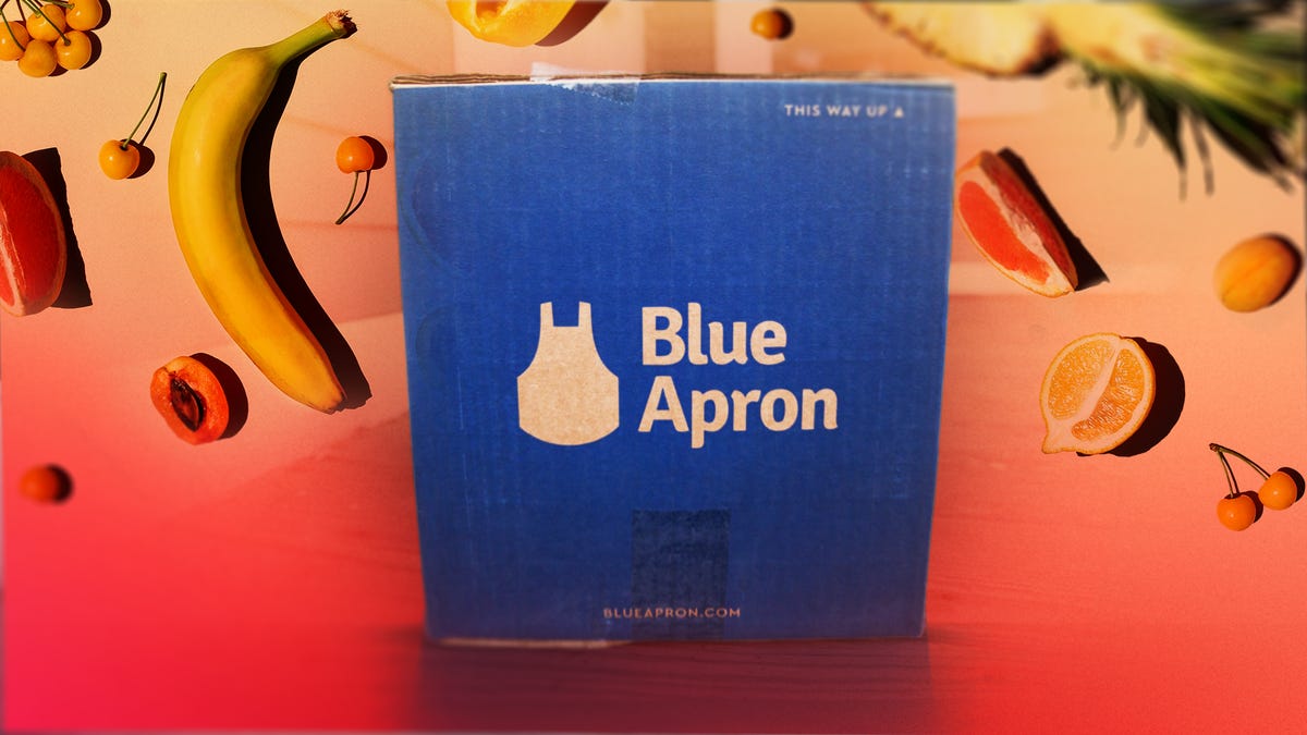 A Blue Apron box against an orange background with fruits behind it.