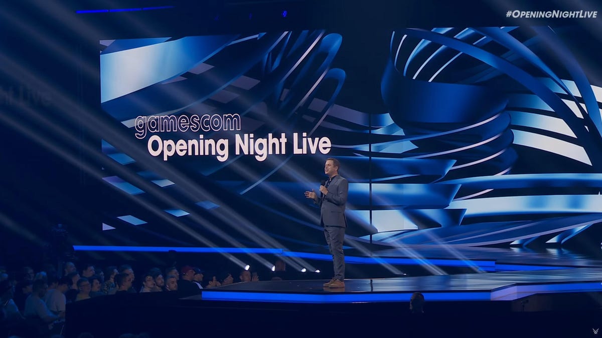 A man stands on stage with a big screen behind him reading "gamescom Opening Night Live"