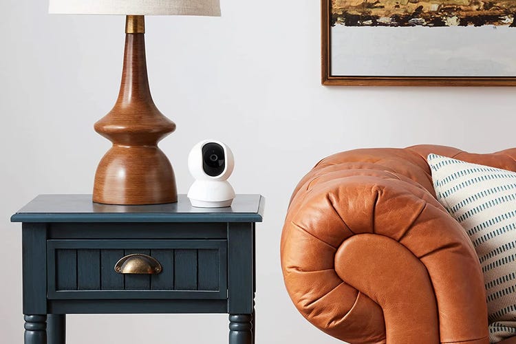 A Tapo pan/tilt cam sits on a dark end table beside a lamp, next to a brown leather couch.