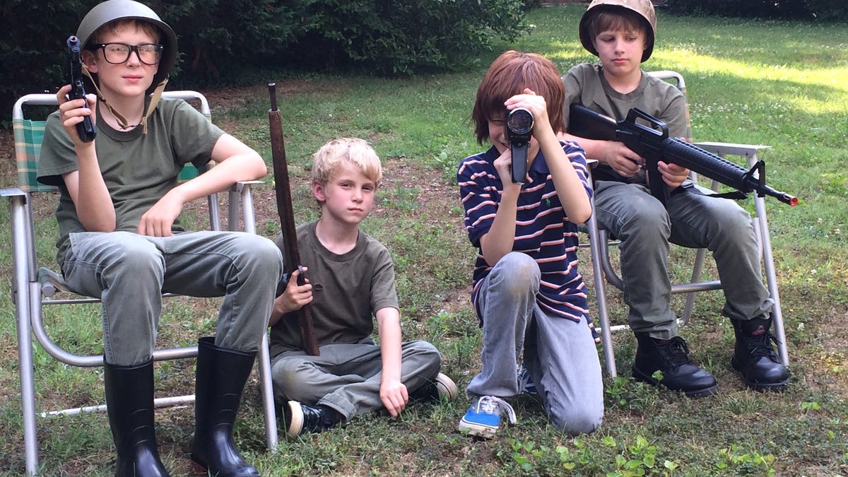 Three boys in olive drab T-shirts, two wearing helmets, hold toy guns. A fourth boy, in a striped Polo shirt, holds a movie camera to his eye.