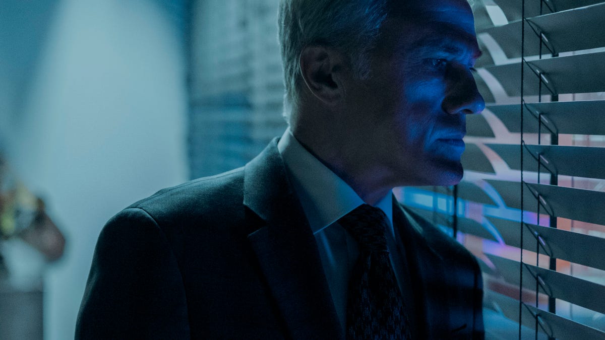 Christoph Waltz in a suit peering through the blinds of an office in shadow