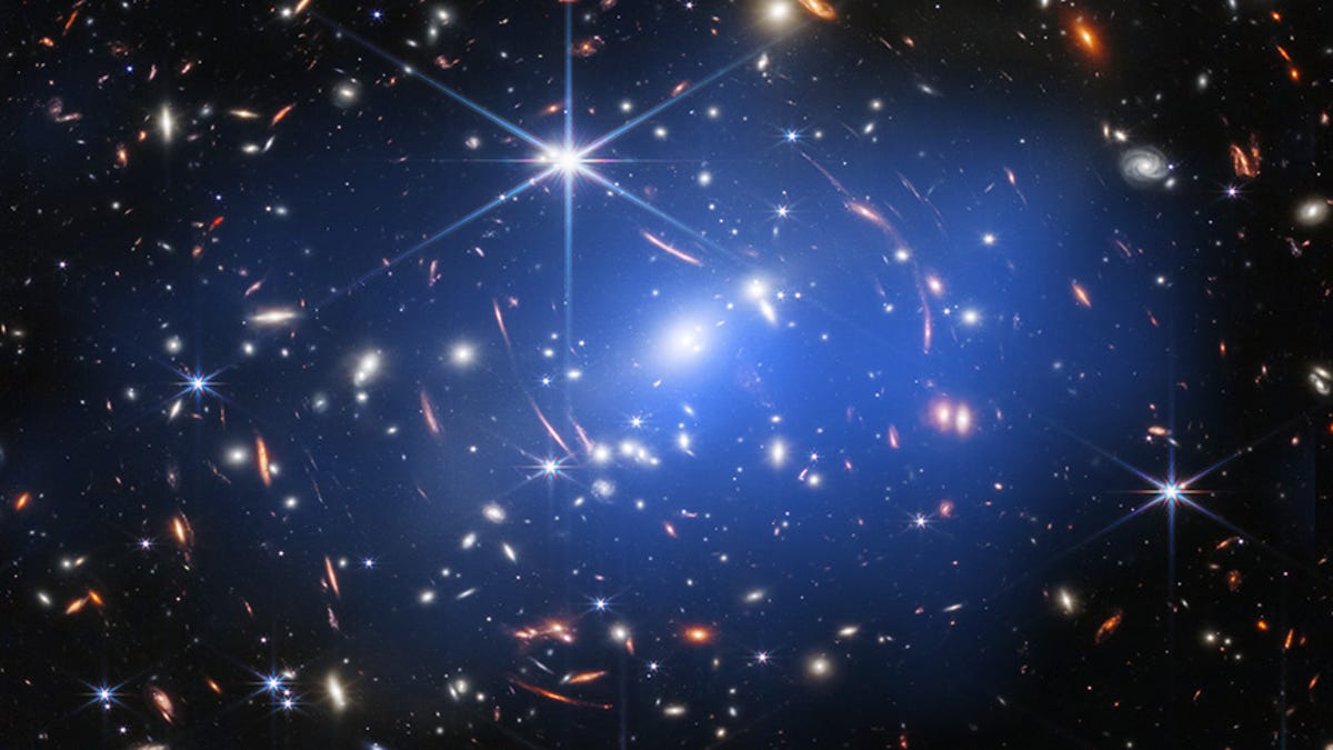 Across the dark canvas of space lie lots of colorful speckles representing galaxies. Many look like streaks because they're warped from our vantage point by gravitational lensing. In the center is a blueish haze representing Chandra observations.