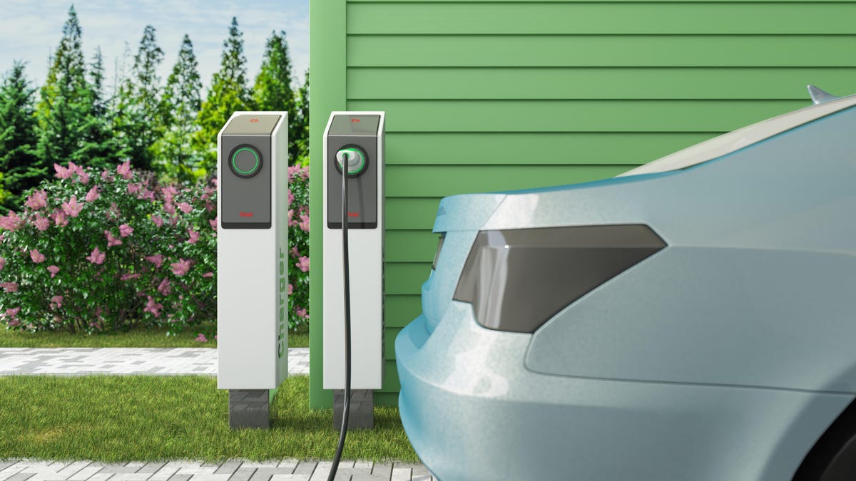 the back of an electric vehicle is shown in front of two charging units, which are in front of a green paneled house sitting next to a yard with flowers and trees