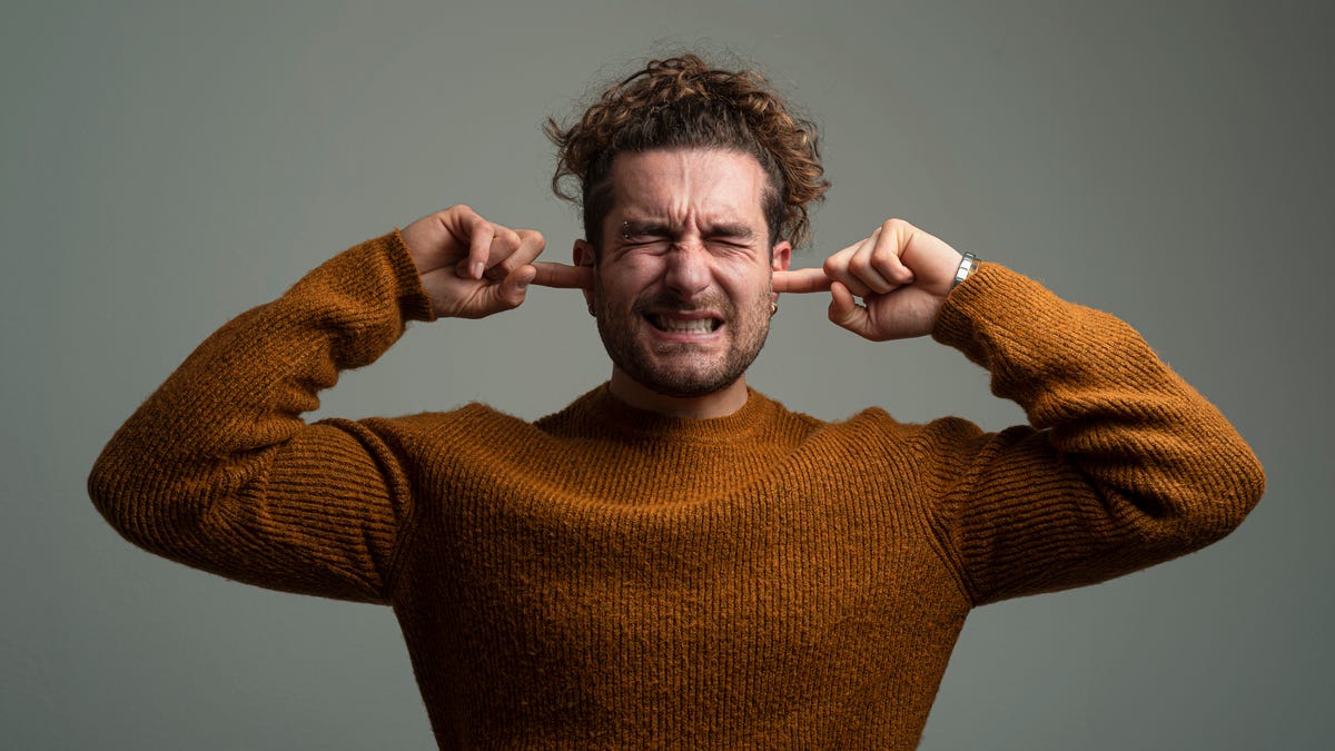 A guy in an orange sweater squinting, plugging his ears