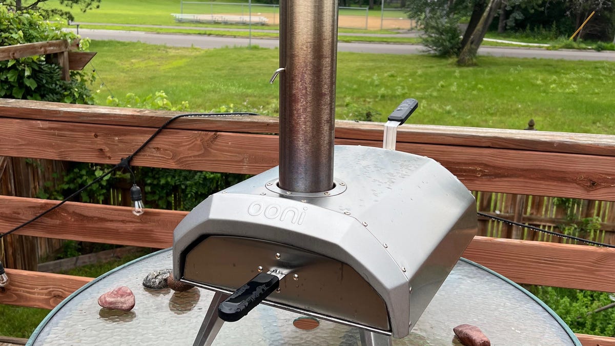 A metal pizza oven sitting atop a round glass tabletop on a wooden deck overlooking a grassy lawn.