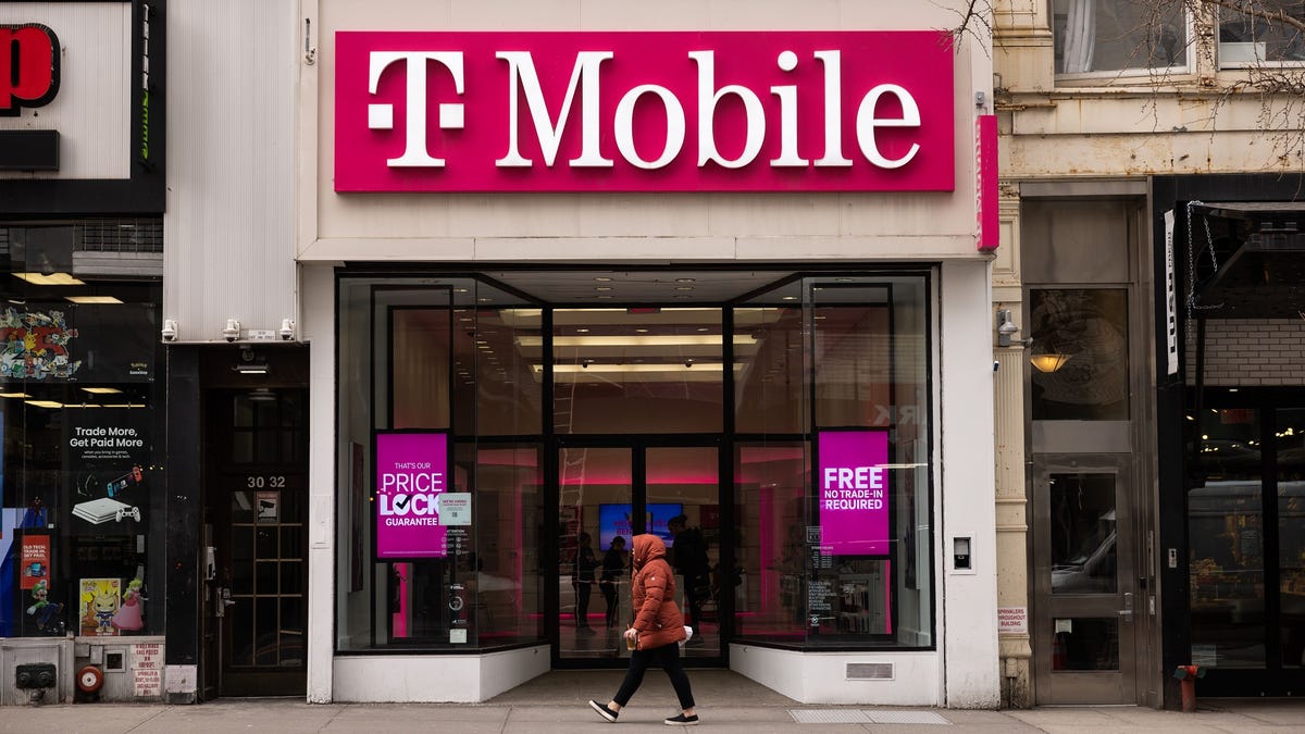 A person in a red jacket walks in front of a T-Mobile storefront on an otherwise empty street.
