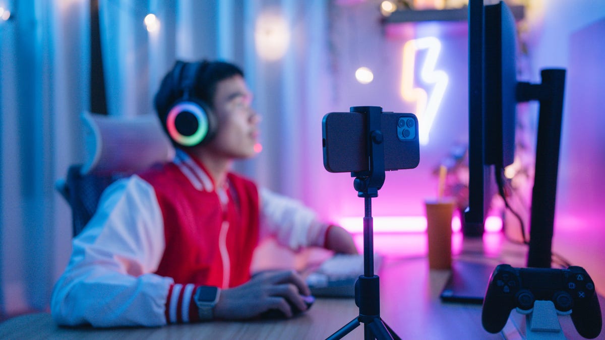 A man sits at a desk in front of a desktop computer wearing rainbow neon headphones while a phone on a small tripod films him