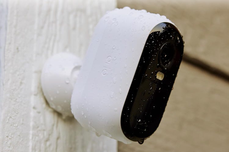 An Arlo Essential cam mounted on white siding and wet with rain.