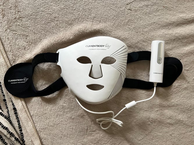 The CurrentBody LED Light Therapy Face Mask on a beige fabric surface.