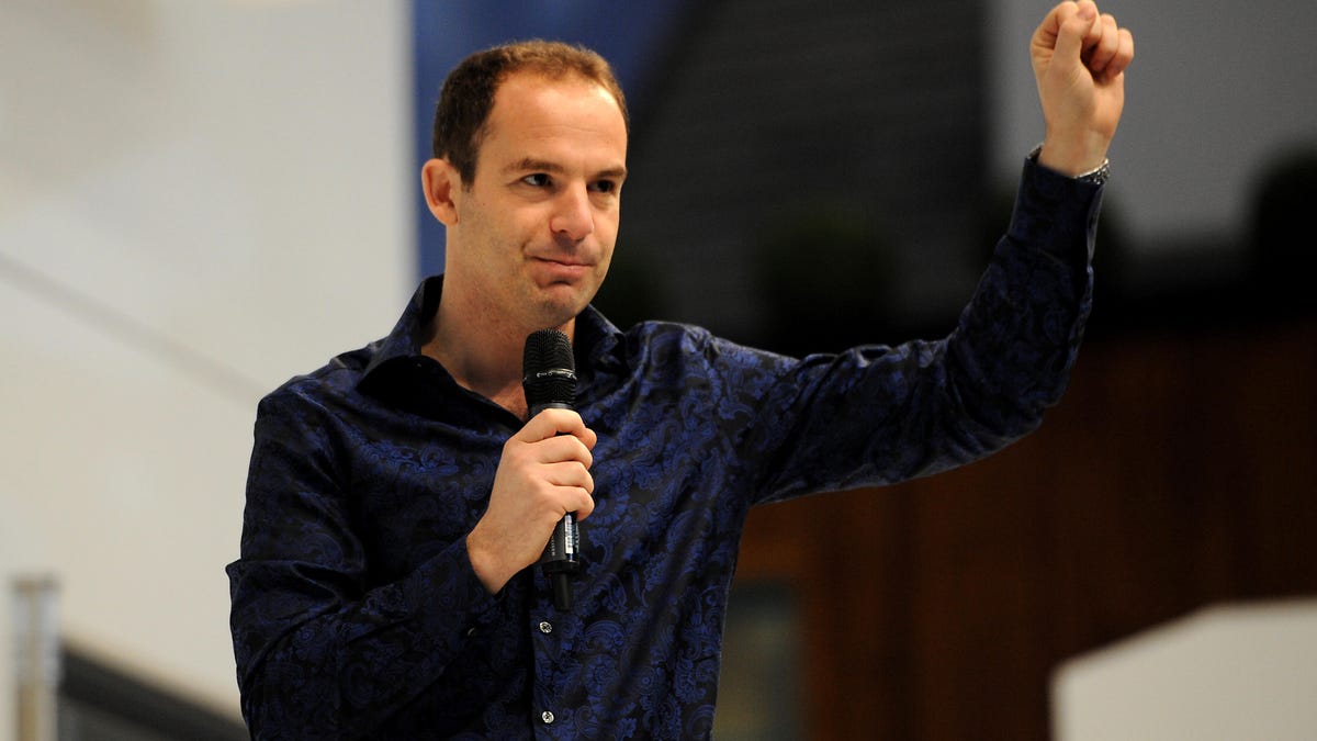 A medium-distance photo of Martin Lewis in a dark blue button down shirt and holding a microphone in his right hand while holding up his right fist.