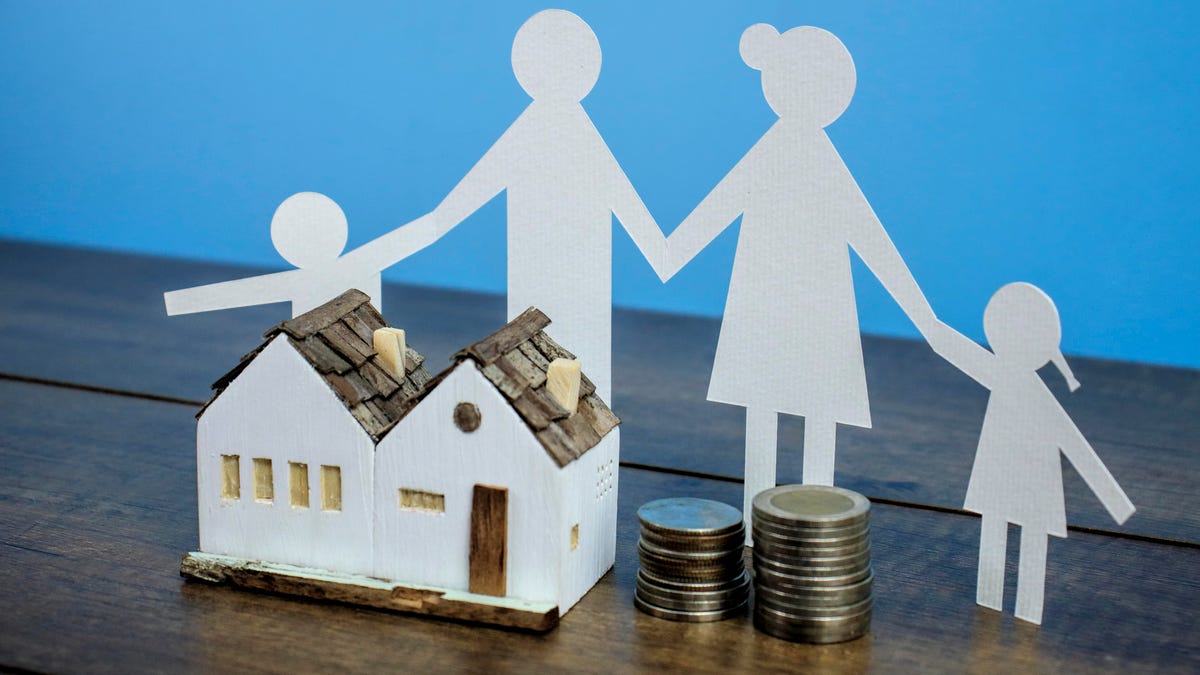 a paper cutout of a family stands behind a small white model house and two stacks of coins