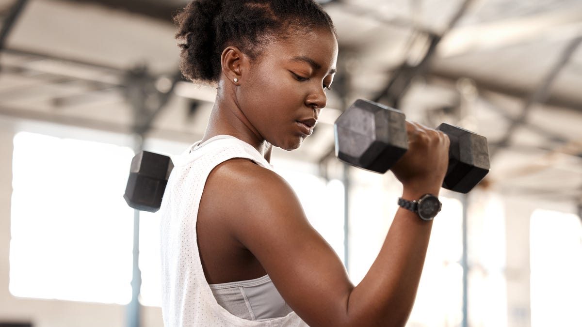 A person with pulled-back brown hair and a white tank top lifting a dumbbell while flexing their muscle.