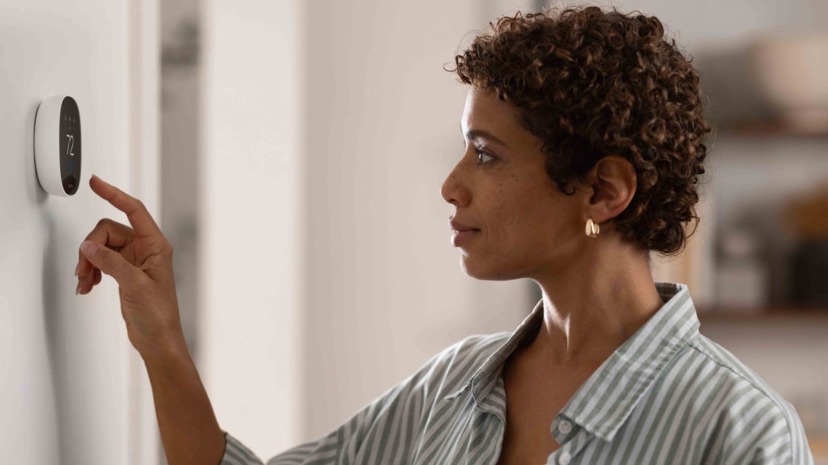 A woman reaches to an Ecobee Essential thermostat on a white wall.