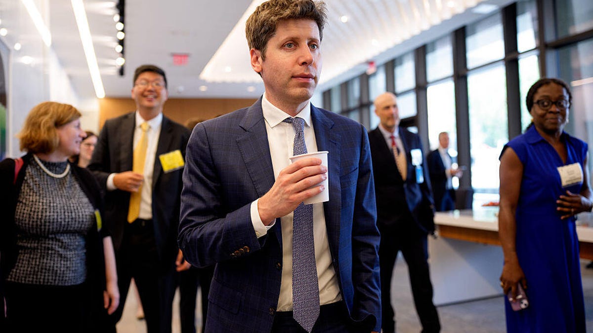 Sam Altman walks in a hallway while holding a cup of coffee.