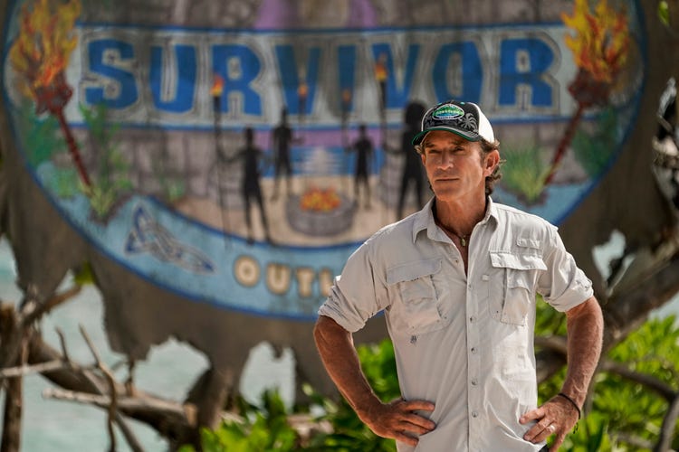 Jeff Probst stands with hands on hips in front of Survivor sign