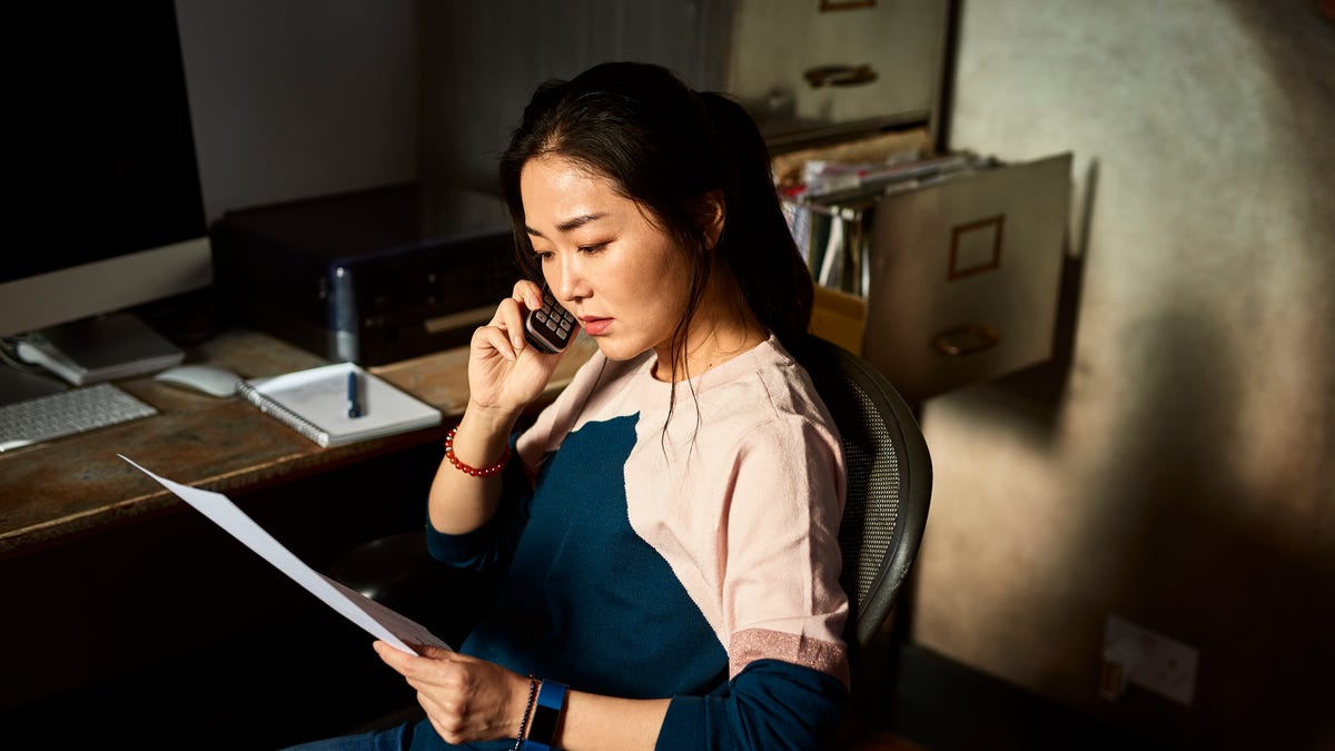 A woman sits in an office chair at a desk while holding a piece of paper and speaking on the phone.