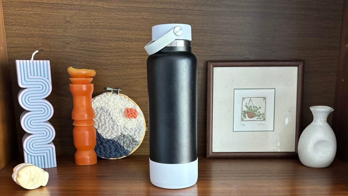 A black Hydro Flask water bottle on a wood cabinet shelf next to sculptural candles, an illustration of a plant, a white vase, and a punch-needle craft.