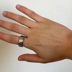 A hand wearing the silver Oura Ring 4 on the pointer finger in front of a white wall.