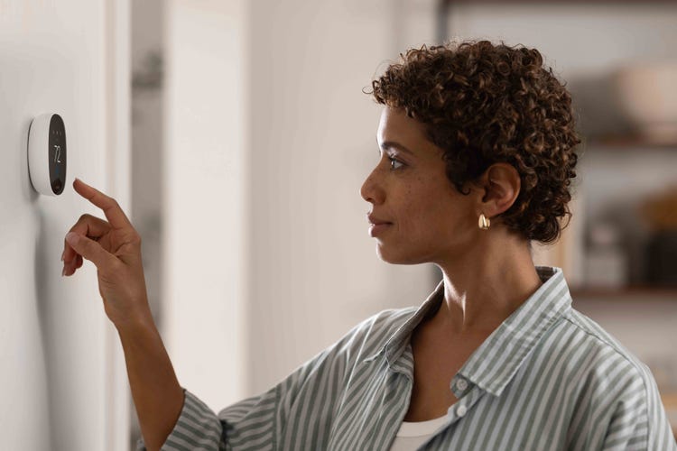 A woman reaches to an Ecobee Essential thermostat on a white wall. 