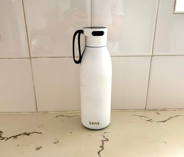 The white Sans Self Cleaning Water Bottle on a white marble countertop with a white tile background.