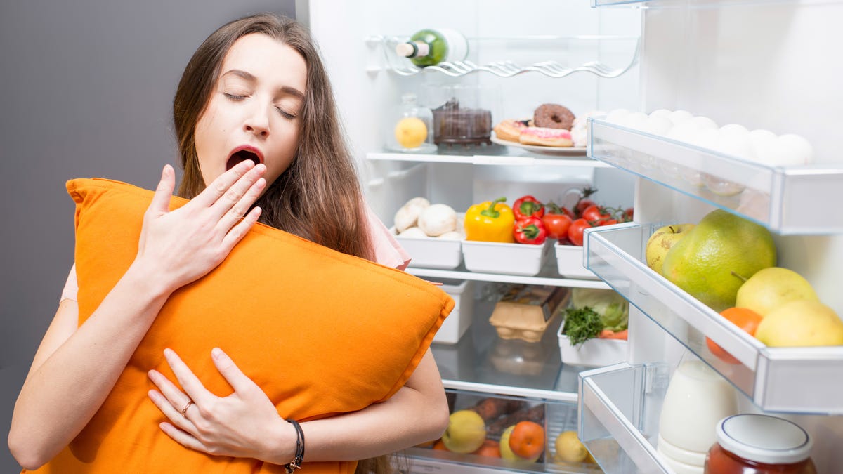 Woman yawns near an open refrigerator while holding a pillow