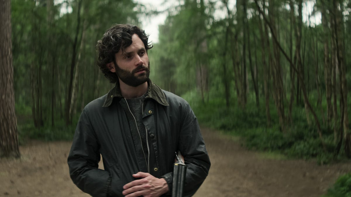 Joe Goldberg standing in a forest with a shot gun over his arm