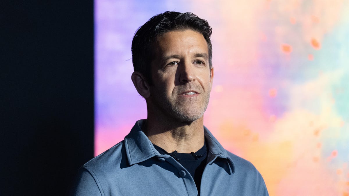 A man with short dark hair and a blue button-down shirt stands in front of a colorful screen and looks off-camera