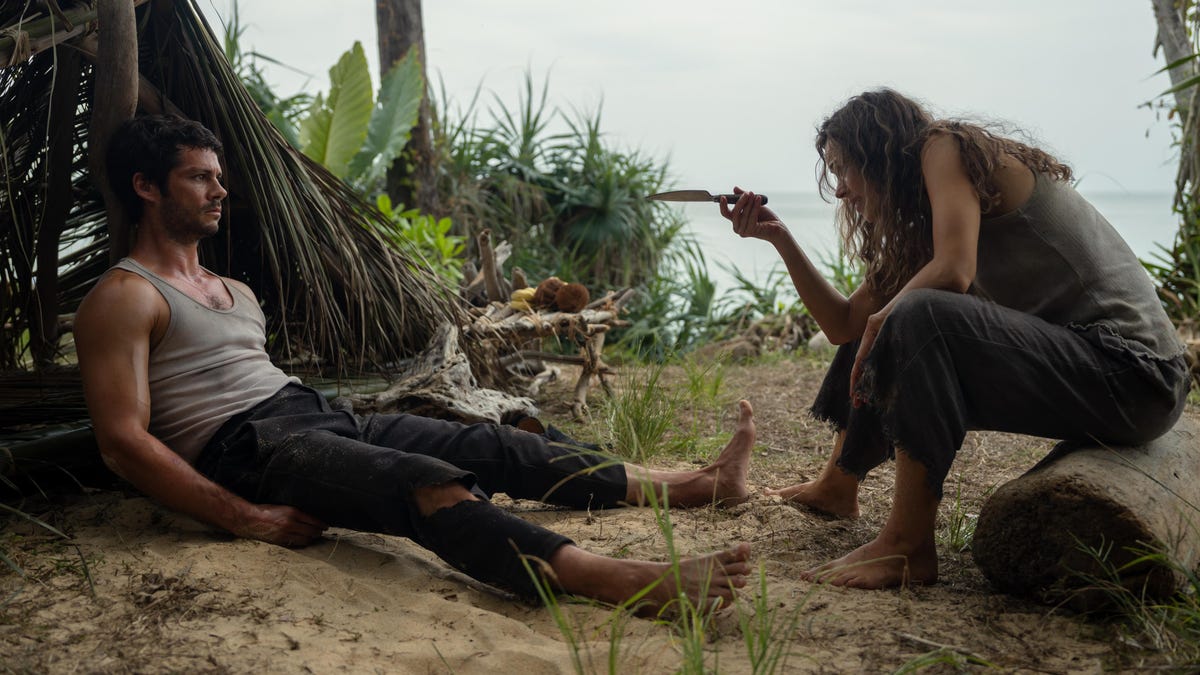 man leans against palm tree as woman sits and talks to him while pointing a knife.