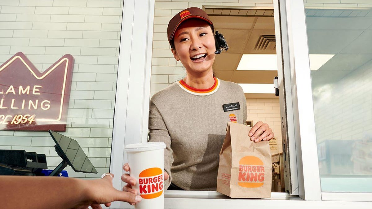 A woman wearing a headset offers a drink and bag of Burger King food to a customer through a drive-thru window.
