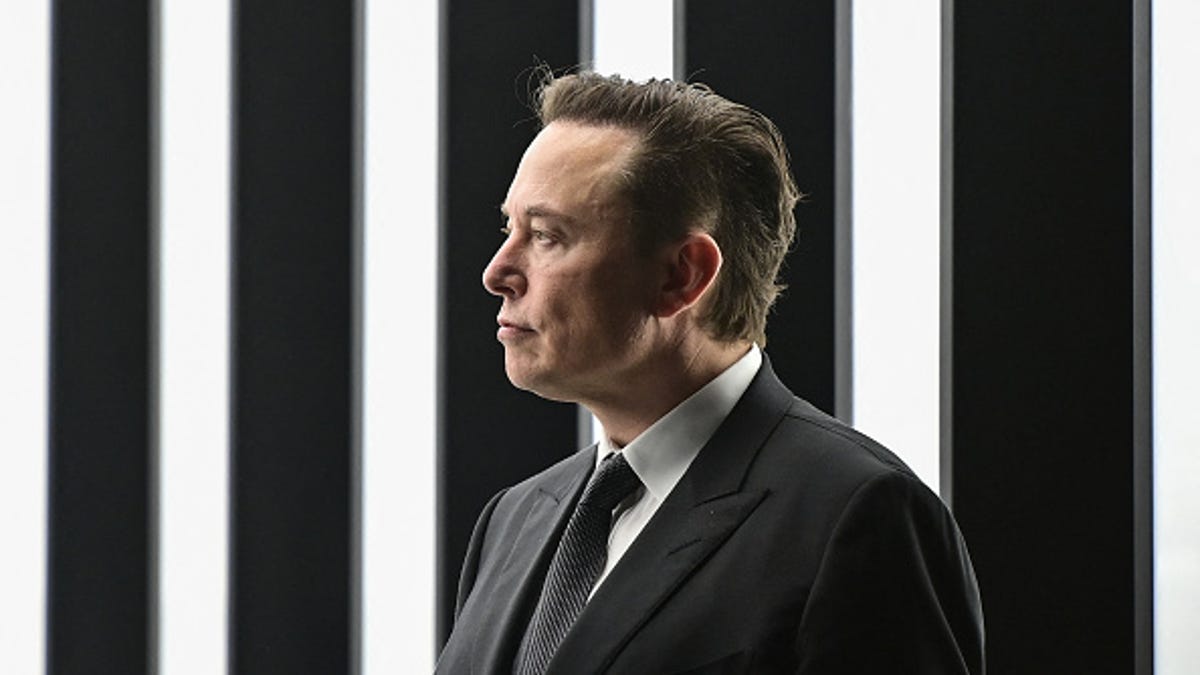 Elon Musk wearing a dark suit and tie, in front of black and white vertical stripes