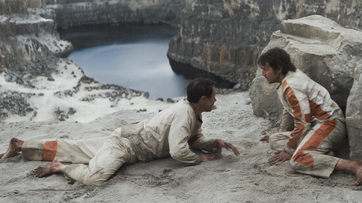 Melchi and Cassian talk as they lie on a sandy cliff overlooking a quarry in Andor