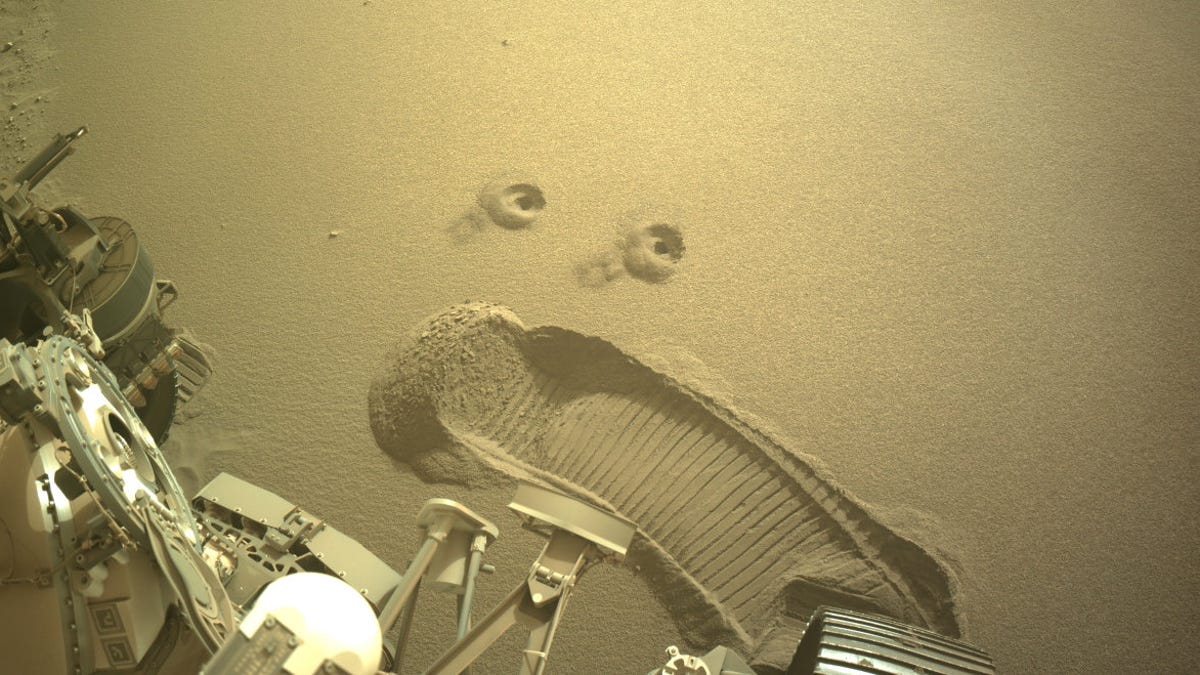 Part of the Perseverance Mars rover is visible at the bottom, but the star is a set of two holes that look like eyes and then a tread scuff that looks like a screaming mouth carved out of the Martian ground.