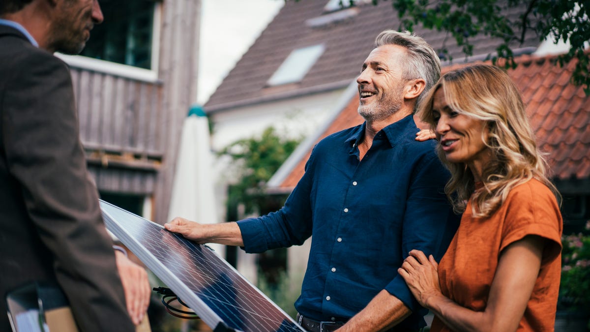 A couple holds a solar panel in front of a home.