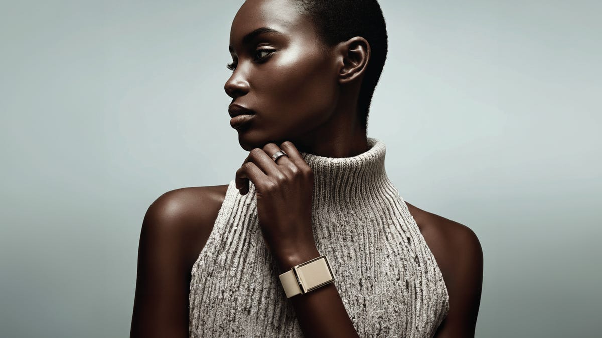 A person in a sleeveless gray turtleneck wears a beige Luna Band. The person is standing in front of a light green backdrop.