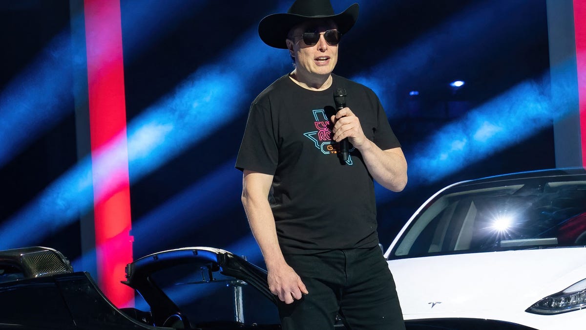 Elon Musk in black T-shirt, sunglasses and cowboy hat, speaking into a handheld mic