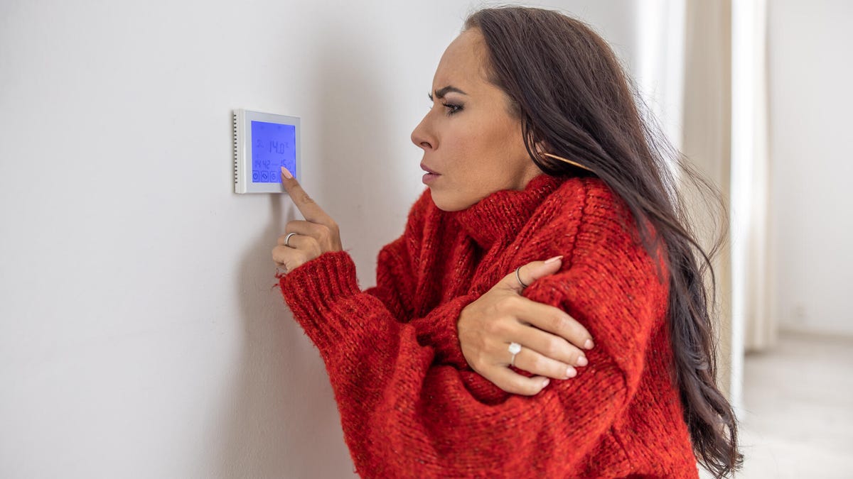 A woman shivering in red adjusts a blue thermostat on a white wall.