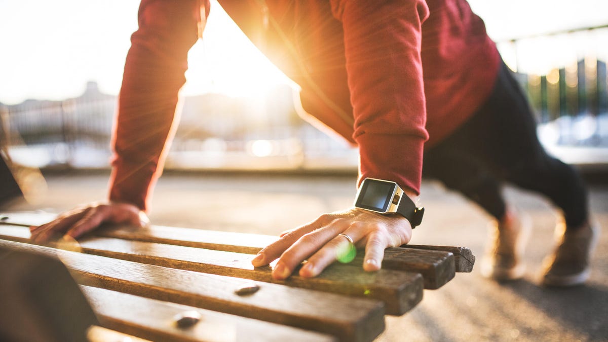 A man in a red shirt with a fitness watch on, doing push-ups on a park bench at sunset.