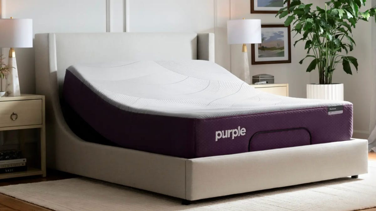 an off-white mattress with purple exterior sitting on light beige upholstered base in a bedroom decorated with an ivory rug and nightstands.