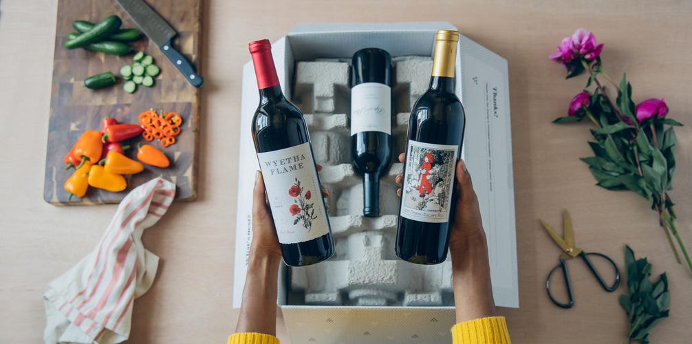 A woman's hands hold two bottles of wine from Firstleaf Wine Club and one more bottle sits in a box in the background.
