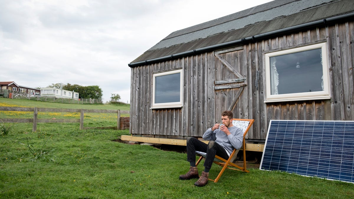 A man sits sipping a cup of coffee in front of a rustic home with a solar panel sitting next to him.